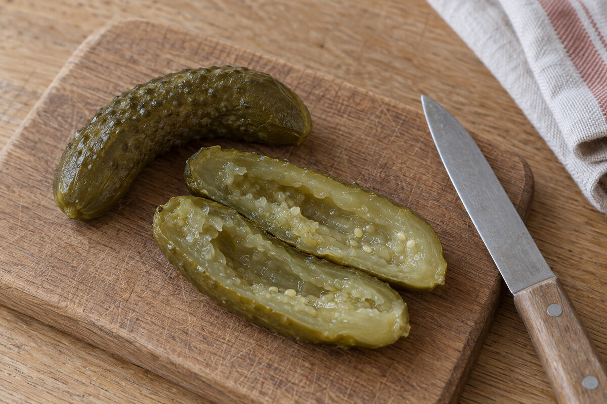 A fermented pickle cut lengthwise, hollow and soft inside, next to a whole one that bends, on an oak board